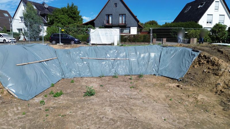 Baugrube mit folienabgedeckter Wand und Bauzaun auf einem Grundstück im Wohngebiet