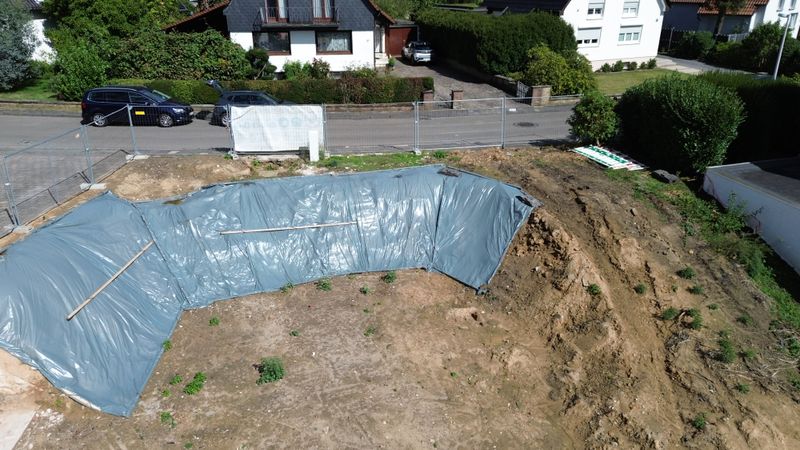 Aushubfläche mit Folienabdeckung und Bauzaun zur Straße, aus der Luft
