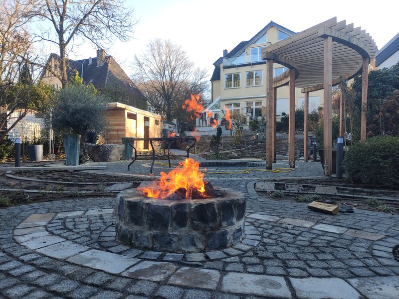 Brennende Feuerstelle aus Naturstein im Pflasterrondell, dahinter Holzpergola, Referenzprojekt Essen